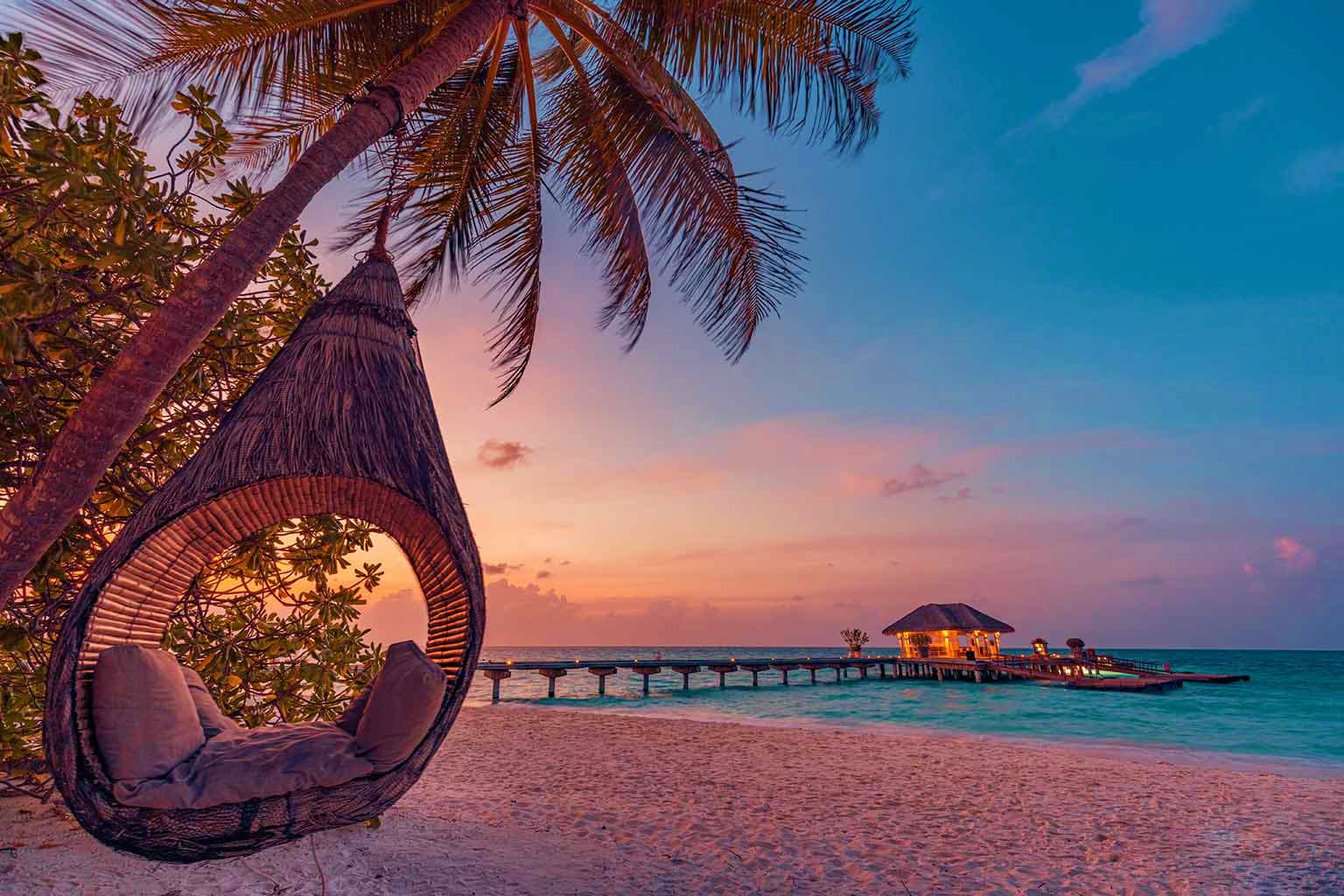 Philippines Visa Hanging woven chair on palm tree overlooking tropical beach with overwater bungalows at sunset in the Maldives